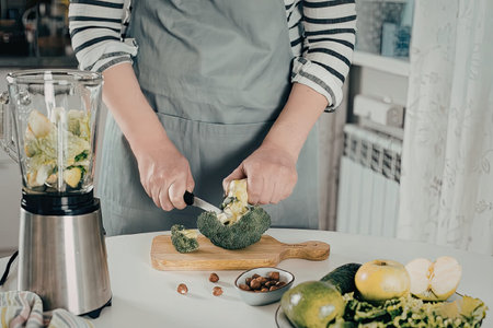 A person wearing a grey apron and a striped shirt is cutting a fresh head of broccoli on a wooden cutting board in a kitchen setting. A blender containing green leafy vegetables sits nearby on the counter, surrounded by various green fruits and vegetables like apples, cucumber, and lime, indicating the preparation of a healthy, nutritious green smoothie.の写真素材