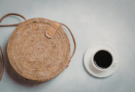 A round woven bag with a leather strap next to a cup of coffee on a white surfaceの写真素材