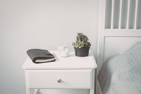 A white bedside table with a single drawer, holding a book, a small potted plant, and a decorative item, next to a bed with a light blue pillow.の写真素材