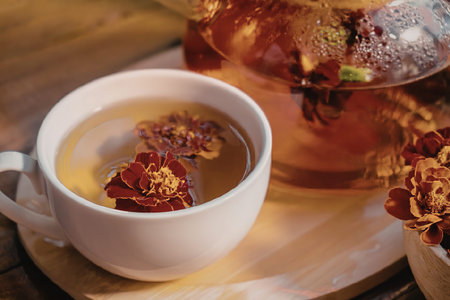 A white cup of herbal tea with flowers on a wooden tray, next to a teapot and a bowl of dried...