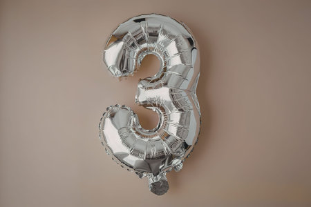 A shiny silver balloon in the shape of the number three against a beige backgroundの写真素材
