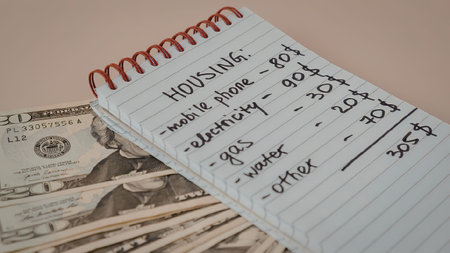 A notebook with handwritten housing bills and expenses on top of a stack of money and matchsticks