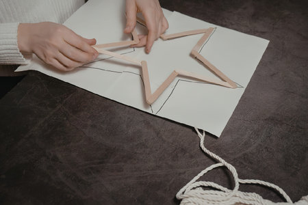 A person is creating a star shaped decoration using wooden sticks and a piece of string on a white...
