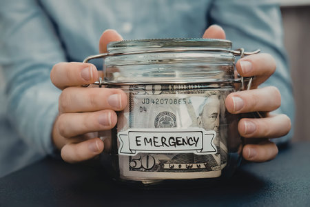 A person holding a glass jar labeled 'EMERGENCY' filled with moneyの写真素材