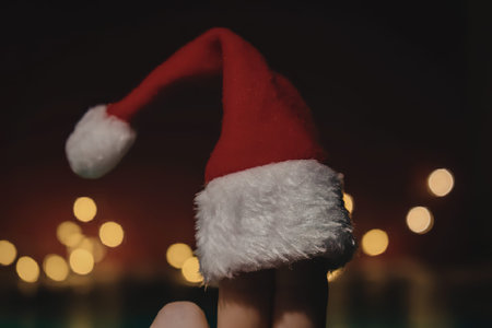 A small red Santa hat with white trim sits on a finger against a blurred background of yellow lights
