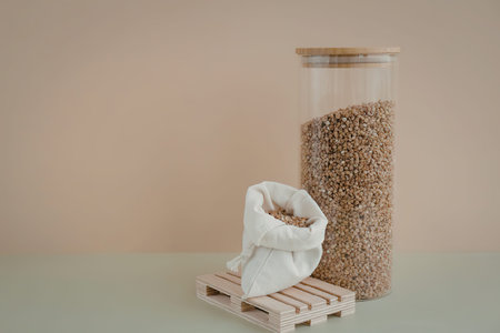A small white sack filled with grain is placed on a tiny wooden pallet next to a tall glass jar...