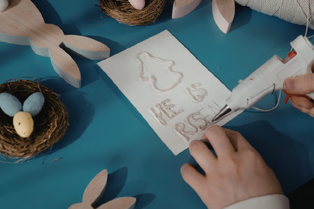 A child's hands holding a hot glue gun and working on an Easter-themed card with the words 'He Is...