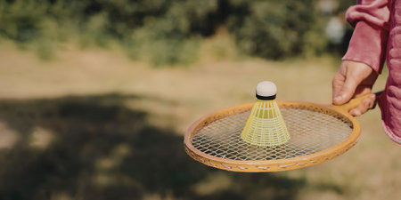 A child's hand holding a badminton racket with a green shuttlecock on it, in a grassy outdoor...