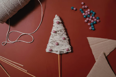 A handmade Christmas tree craft made from yarn and beads on a stick, surrounded by crafting...