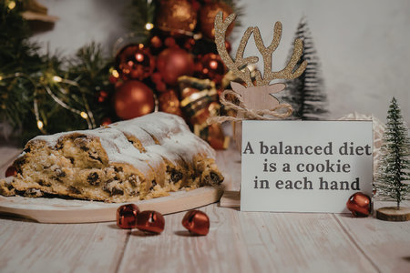 A festive Christmas stollen with icing on a wooden plate, surrounded by red ornaments, small Christmas trees, and a sign that reads 'A balanced diet is a cookie in each hand'.の写真素材