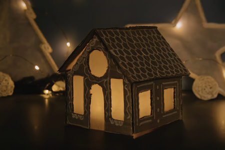 A warmly lit gingerbread house with glowing windows sits on a dark surface, surrounded by snow and lights.の写真素材