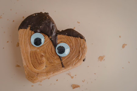 A heart shaped pastry with chocolate on top and googly eyes, on a beige surfaceの写真素材