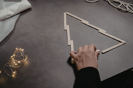 A person's hand assembling a wooden Christmas tree on a gray surface with string lights nearby.