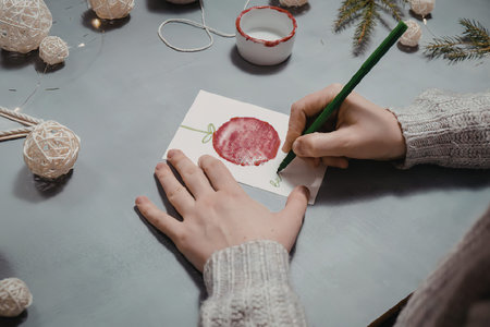 A person painting a red circle on a white card with a green brush on a table with Christmas...