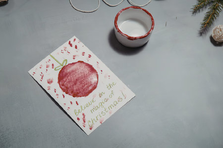 A handmade Christmas card featuring a red apple on a white background with pink and green designs,...
