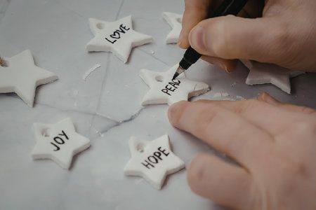 A person's hand is shown writing on white star-shaped ornaments with a black marker, with words like...