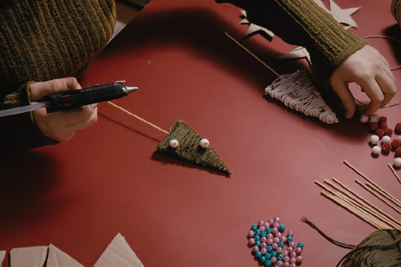 A person crafting a small felt Christmas tree with beads and string on a red table.