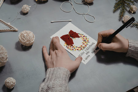 A person's hands are shown decorating a Christmas card with a red bow and yellow flowers on a gray...