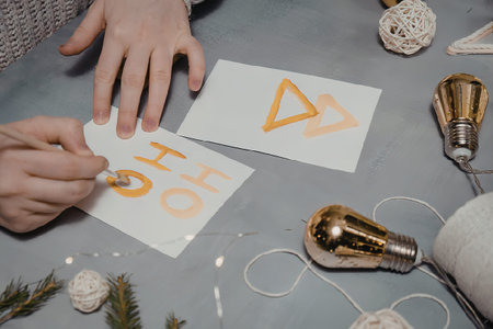 A person's hands are shown decorating Christmas cards with orange cut-out letters and shapes on a gray table with lights and ornaments.の写真素材