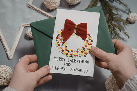 A person holding a Christmas card with a green envelope, featuring a red bow and colorful wreath...