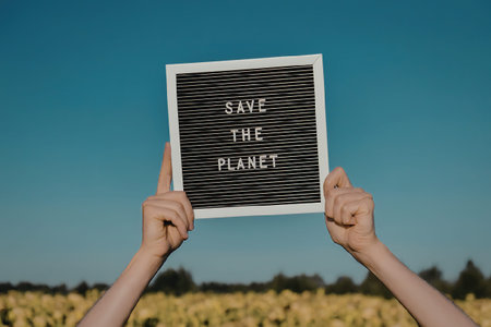 A pair of hands holding a sign that reads 'Save The Planet' in a field with a clear blue sky.
