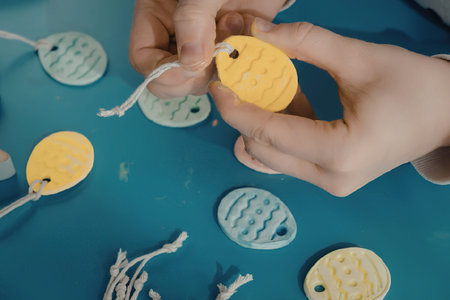 A person's hands holding a yellow Easter egg decoration with a string, surrounded by other colorful egg decorations on a blue surface.の写真素材