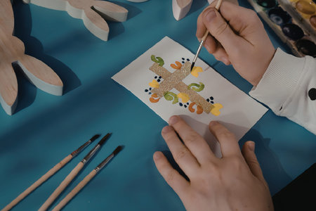 A person painting a vibrant design on a white paper with a small brush, surrounded by paintbrushes and wooden cutouts on a blue surface.の写真素材