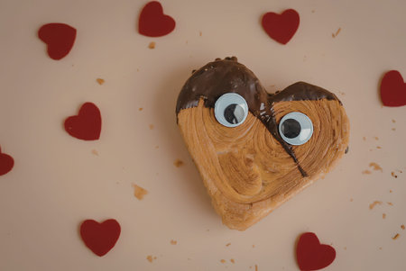 A heart shaped pastry with candy eyes and chocolate on top, surrounded by small red hearts.の写真素材