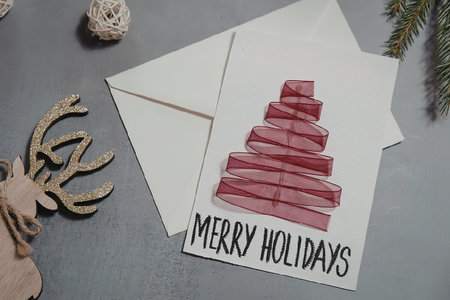 A holiday card with a red Christmas tree design and the words 'Merry Holidays' on a gray surface with a white envelope and holiday decorations.の写真素材