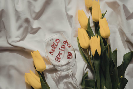 A wine glass with 'Hello Spring' written on it, surrounded by yellow tulips on a white cloth background.の写真素材
