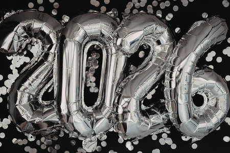 A shiny silver balloon displaying the year 2026, surrounded by confetti on a black background,...