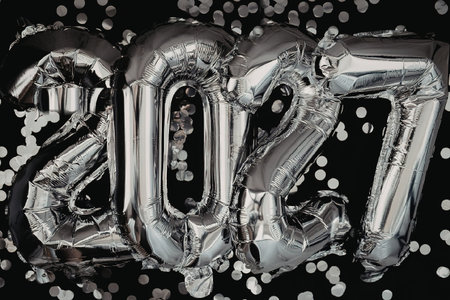 A close-up of silver balloons forming the year surrounded by confetti on a black background, symbolizing celebration and festivity.の写真素材