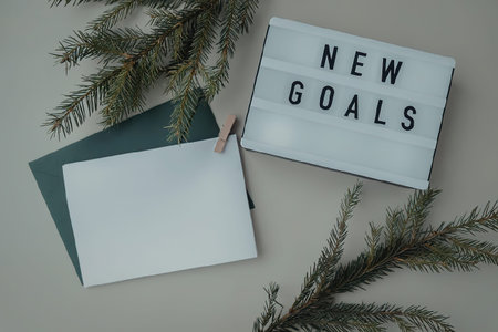 A flat lay image featuring a lightbox with 'NEW GOALS' text, a blank white card, a green envelope, and evergreen branches on a grey background.の写真素材