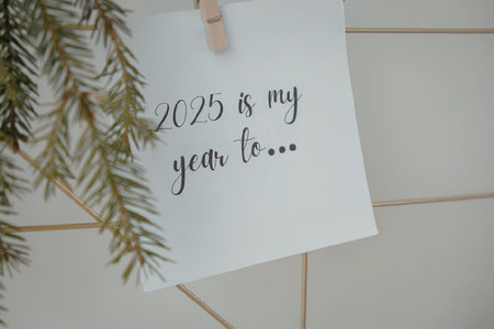 A white card with the text '2025 is my year to...' hangs from a clothespin on a white tiled wall, accompanied by a green pine branch.の写真素材