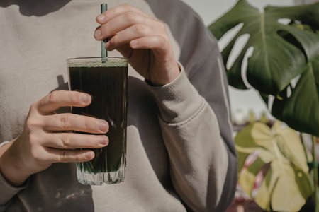 A person is holding a glass of green juice with a straw, standing in front of a plant.