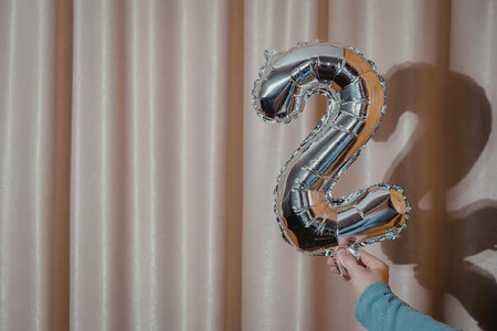 A hand holding a large silver number two balloon in front of a beige curtain.