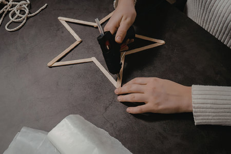 a person's hand holding a hot glue gun and assembling a star shape made of wooden sticks on a tableの写真素材