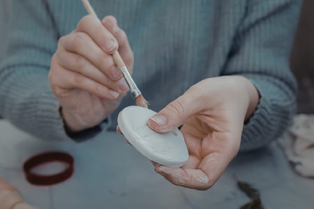 A person wearing a blue sweater is painting a small white object with a brush.