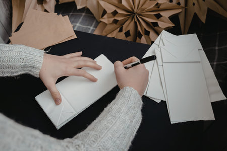 A person is sitting at a table, writing on a piece of paper with a pen. The person is wearing a gray sweater and has their hands visible. There are craft supplies in the background, including paper snowflakes and a brown paper bag.の写真素材