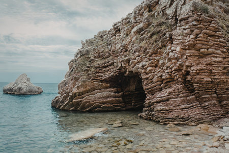 A large rocky cliffside with a cave entrance stands by the calm sea water, with another rock formation visible in the background under a cloudy sky.の写真素材