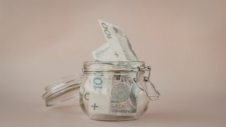 A glass jar with a handle and latch, containing rolled up banknotes, likely euros, on a beige...