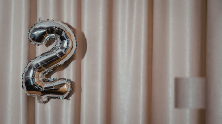 A shiny silver balloon in the shape of the number 2 against a beige curtain backgroundの写真素材