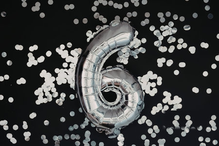 A silver balloon in the shape of the number six surrounded by confetti on a black background.