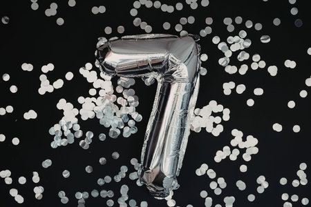 A shiny silver balloon in the shape of the number seven, surrounded by small white confetti on a...