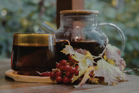 A glass teapot and cup filled with dark tea, accompanied by red berries and leaves on a wooden...