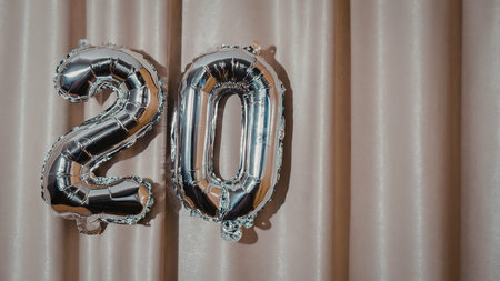 A close-up image of silver balloons forming the number 20 in front of a beige curtain, likely used...