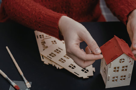 A person in a red sweater assembling a small wooden house with a red roof on a table with paintbrushes.の写真素材