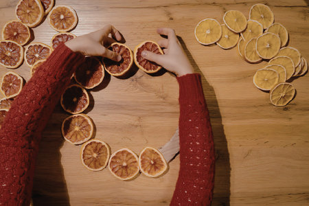 A person in a red sweater making an orange wreath on a wooden tableの写真素材