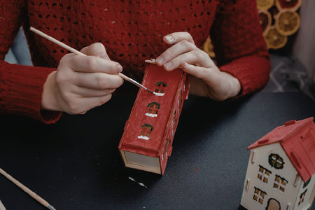 A person in a red sweater painting a miniature red house model with a small brush.