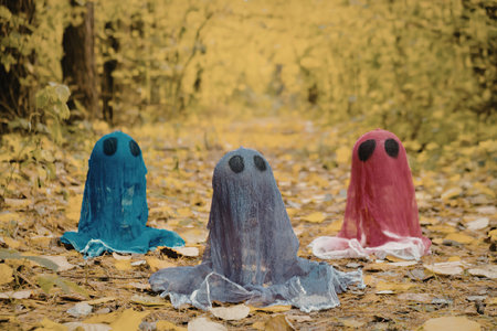 Three ghostly figures made of colored fabric sit on a forest floor covered with autumn leaves. The...